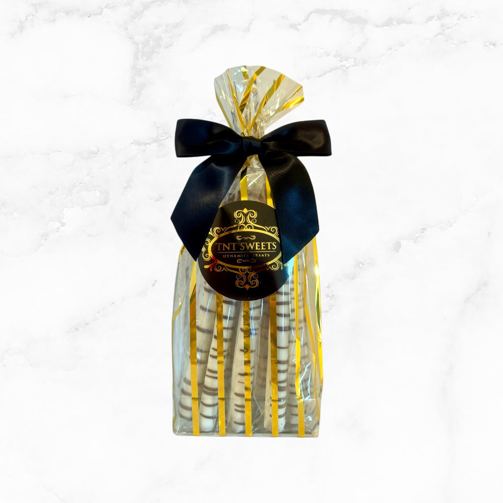 White chocolate dipped pretzels in a gold striped bag with a black bow and label on a marble countertop.