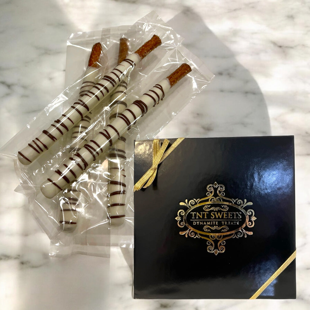White chocolate dipped pretzels next to a TNT Sweets black and gold gift box on a marble countertop.