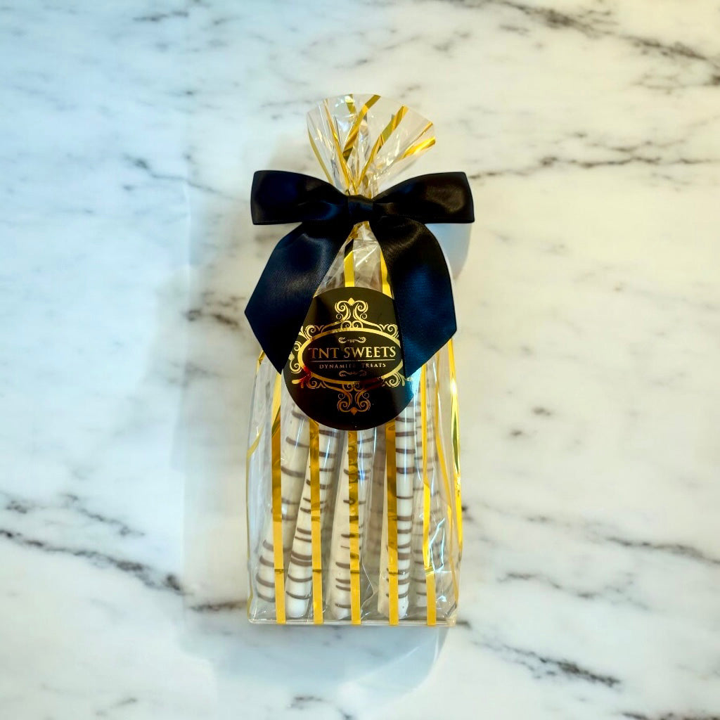 White chocolate dipped pretzels in a gold striped bag with a black bow and label on a marble countertop.